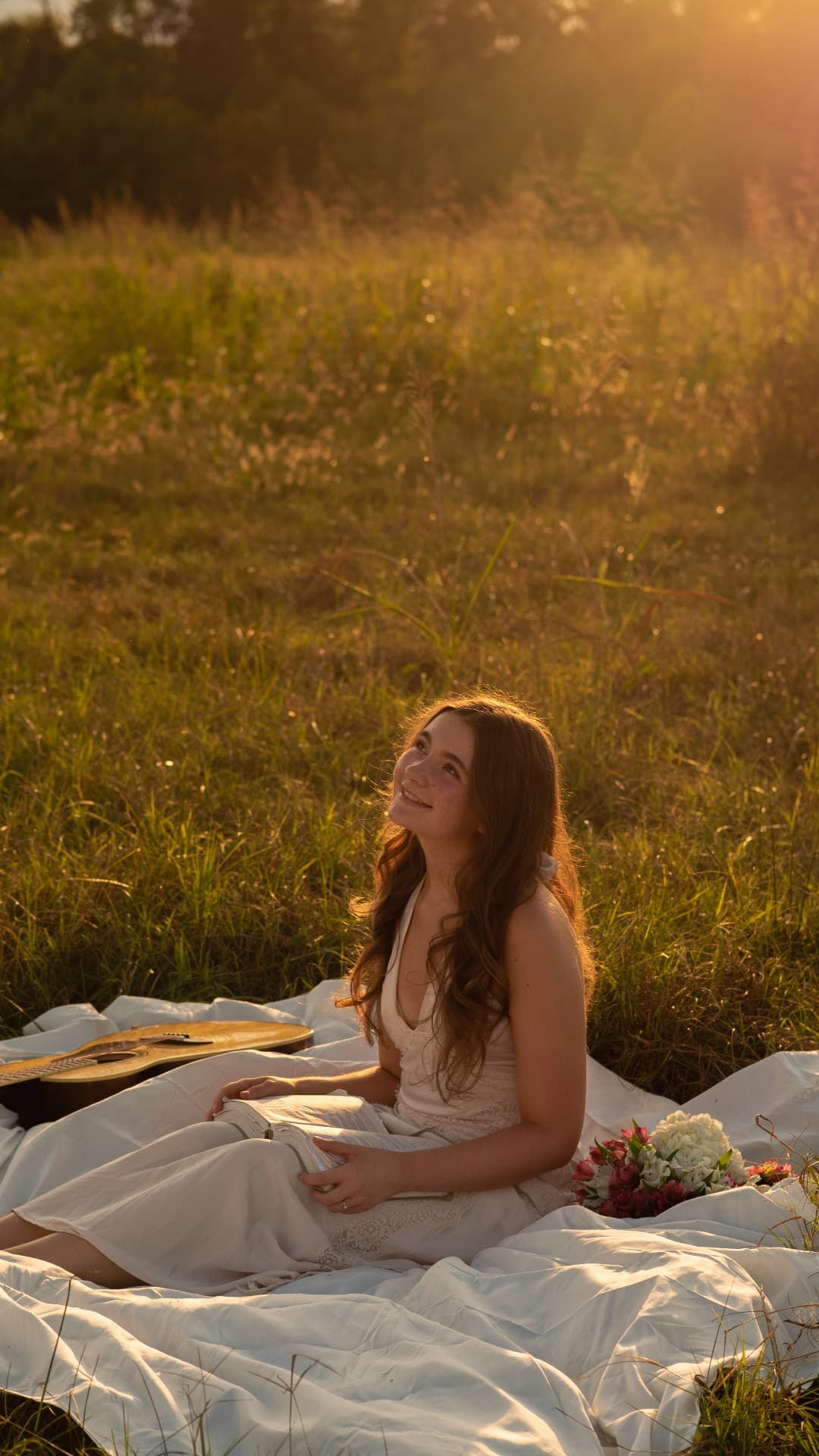 A girl laughing on a white blanket with her guitar and a book in the evening light