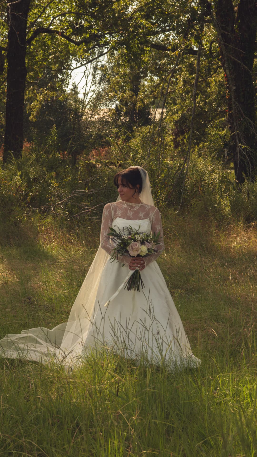A bride standing in a sunlit forest clearing, veil trailing through the grass