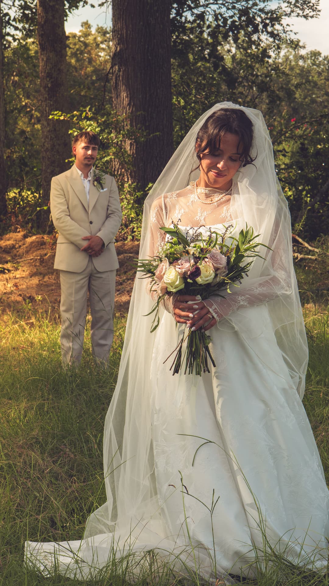 A bride looking down at her bouquet while the groom waits behind